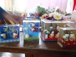 机の上に、透明な箱に収められた4つの日本の伝統的な人形や、花束などのまゆ細工が並べられている写真