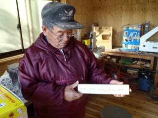 屋内の作業場で、赤いジャンパーを着た帽子の男性が白い長方形の木材の商品を手に持っている写真