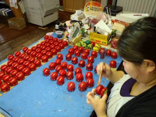 机の上に並べられた多数の赤いタコ型の文鎮が並び、女性が一つを手に取り丁寧に色付けをしている写真