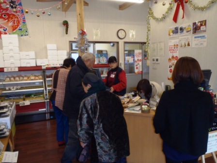 棚に商品が並ぶ店内で、複数の買い物客がレジカウンターに並び、店員が対応している様子の写真