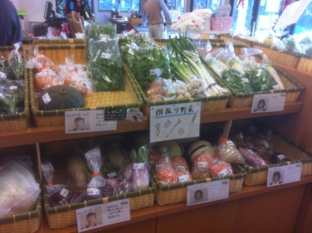 かごに入ったネギやほうれん草、ニンジン、カボチャなどの新鮮な野菜が棚に並べられ、それぞれの野菜のそばには生産者の顔写真と名前が添えられている、野菜コーナーの写真
