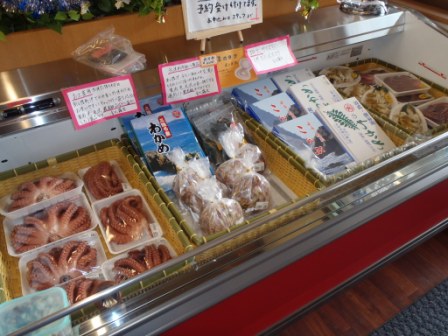 店内の冷蔵ケースにゆでダコやワカメ、冷凍ホヤや海産物加工品などが丁寧に並べられ、上部には手書きの説明ポップが掲示されている、海産物販売コーナーの写真