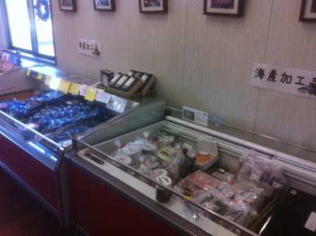 店内の壁際に沿って設置された冷凍ケースに、冷凍魚や海産加工品が種類ごとに分けて整然と並べられ、壁には「鮮魚コーナー」や「海産加工品」といった表示が貼られている写真