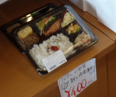 梅干しが乗った白ご飯、揚げ物、卵焼き、焼き魚、サラダなどのおかずが色とりどりに詰められた弁当が置かれた棚に、手書きの価格表示と説明文が添えられている写真