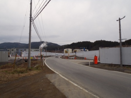 緩やかに曲がる舗装道路の右側には白い仮囲いの塀が並び、奥に災害廃棄物処理施設の白いテントや重機が見える写真