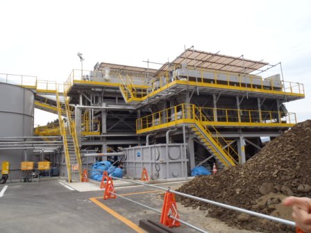 階段や手すりが黄色で縁取られた三階建ての大型の機械設備が中央にあり、右手前には大きな土の山、左奥には大型タンクが見える土砂の洗浄設備の写真