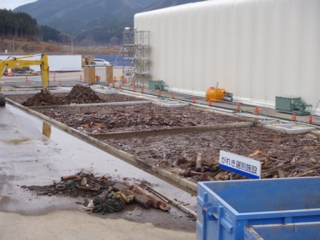 がれき選別施設と書かれた看板があり、3つの区画で仕切られたプールに、木材などのがれきが浸かっている様子の写真