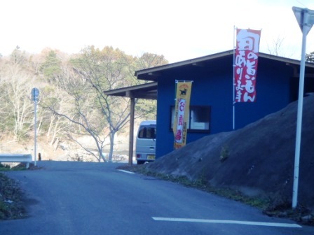 のぼり旗が立つ小道を進んだ先の右手に、黒い外壁の平屋建て事務所が見えている様子を写した写真