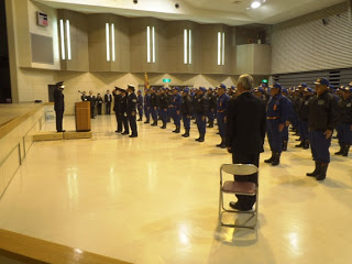 広い屋内ホールで、消防団と交通指導隊と思われる制服姿の人々が整列しており、代表の関係者が壇上に立つ男性と向かいあい、敬礼をしている出初式の様子の写真