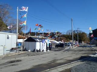 青空の下、伊里前福幸商店街の道路の両側の店先に色とりどりのJリーグフラッグが立てられ、旗が風になびいている通りの様子を写した写真