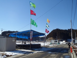 雪が積もった道路の脇に青いシートで屋根がわれた建物があり、建物の柱に3枚づつたJリーグのフラッグが掲げられている様子が写っている写真