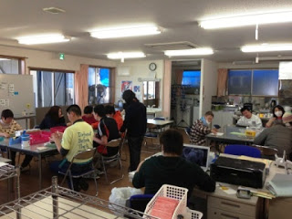 室内の作業場でたくさんの人がグループに分かれて机に向かって作業を行っている作業風景の写真