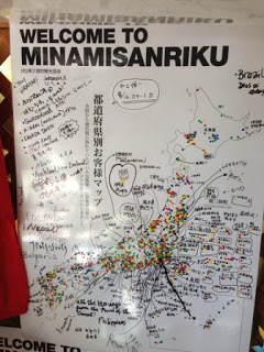 「WELCOME TO MINAMISANRIKU」と書かれた白地のポスターで、中央に日本地図が描かれ、その周囲にメッセージが手書きで記され、各地からの訪問者が自分の出身地に色とりどりの印をつけているポスターの写真