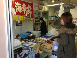 魚屋の店内で、カウンターに魚や海産物が並べられ、奥では店員の男性が作業をしており、手前には女性の買い物客が来店している店舗内の様子を写した写真