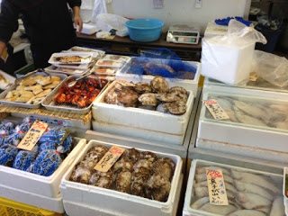 白い発泡スチロールの箱に貝や魚などの魚介類が並べられ、奥には焼き物や青いネットに入った品が並んでいる様子の写真