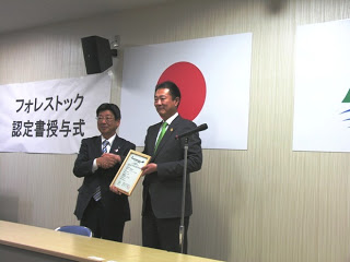 日本国旗とその横にフォレストック認定書授与式と掲示され、スーツ姿の男性2名が額に入ったフォレストック認定書を手に持ち笑顔で映っている授賞式の様子の写真