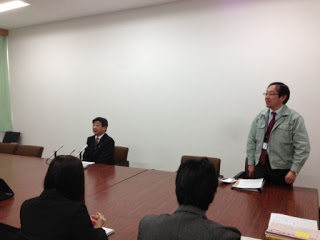 記者たちを前に、町長が記者会見のために席に着き、隣で職員の男性が立って話をしている様子を写した写真