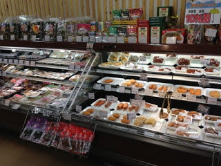 陳列棚にパック詰めされた総菜など様々な食品が並び、上段には調味料や乾物などの商品が陳列されている店舗内の様子の写真