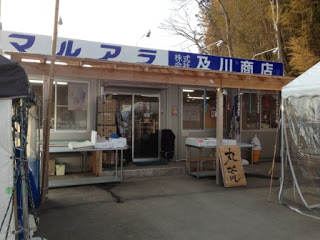 「マルアラ 及川商店」と書かれた看板が掲げられた木造の屋根の商店の前に、商品が並べられたテーブルと「丸荒」の立て看板が置かれている写真