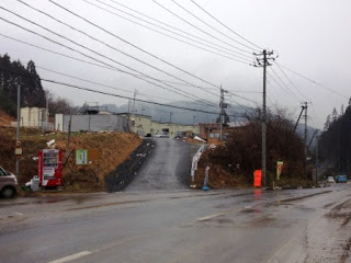 2車線道路の左に仮設住宅に続く坂道があり、坂の上に建物が見える仮設住宅入口の写真