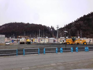 工事用バリケードで仕切られた中に、重機やトラックが停まった整備中の広い空き地があり、その先には仮設商店街の店舗が並んでいる写真