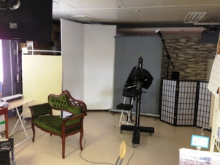 白とグレーの背景スクリーンが設置され、中央には大型カメラと照明機材、左側に緑色のソファ、右奥には和風の仕切りがある室内で、撮影用のセットが整えられたスタジオの写真