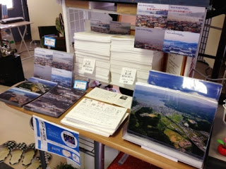 室内の棚の上に、震災前後の南三陸町の様子を写した写真や写真集が整然と並べられており、棚の下には青いチラシが貼られた展示コーナーの写真