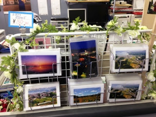 白い金属製のラックに風景写真のポストカードが6種類並べて掛けられ、周囲は白い造花のガーランドで飾られたポストカード販売コーナーの写真