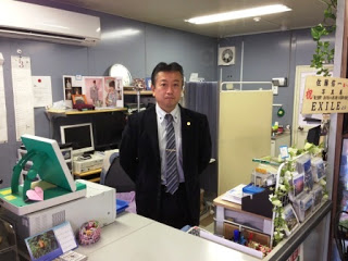 スーツ姿の佐藤さんが佐良スタジオ受付のカウンター内に立って写っている写真
