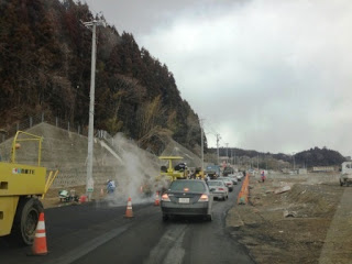 工事が進められている道路で、アスファルトを舗装する大型機械や作業員たちの姿があり、その横では車が一列に並んで通行を待っている道路工事中の写真