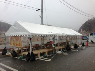 駐車場の敷地に白いテントが設置され、内部には木製のテーブルとベンチが並び、テントの周囲は透明のビニールシートで囲まれている「楽テント福幸」の外観写真