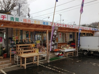 商店の前に野菜や果物、木製のテーブルとベンチなども並んでおり、店舗前が駐車場になっているマルタケ大衆ストアの外観写真