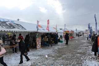 雪が降る中、屋外の市場で白いテントがいくつも並び、その間を多くの人々が行き交いながら買い物をしている賑わいが感じられる市場の写真
