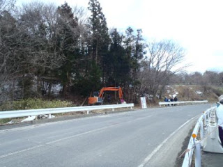 カーブした車道の左側に林が広がり、その手前でオレンジ色のショベルカーが作業しており、右側の歩道には数人の人が集まって様子を見ている工事現場の写真