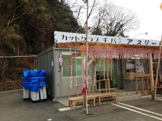 木々が茂る山を背景に、仮設の理髪店が建てられており、店舗の前にはベンチと椅子が並べられている、仮設店舗カットクラブチバの写真