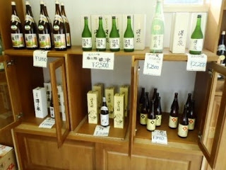 木製の棚に複数の緑や茶色の日本酒の瓶と箱がきれいに並べられていて、それぞれに値札が付けられている店内の写真