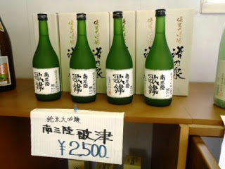 木製の棚の上に、「南三陸歌津」と書かれた白いラベルが貼られた緑色の日本酒の瓶が4本並び、背後には白い箱が並べて置かれ、棚の前面には「純米大吟醸 南三陸歌津 2500円」と書かれた紙が貼られている写真