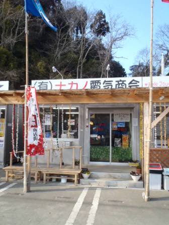 プレハブ風の仮設店舗で、有限会社ナカノ電気商会の看板が掲げられ、店舗の前には旗が立てられているナカノ電気商会の外観写真