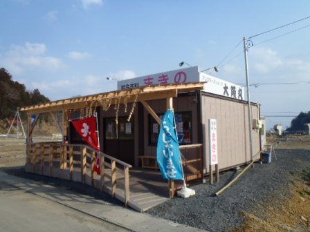 屋根に正面に「まきの」側面に「大隆丸」と書かれた看板が掲げられ、建物の前には赤と青ののぼり旗が立てられている店舗の外観写真