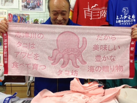 店主の千葉さんが「志津川のタコはアワビを食べて育つ だから美味しい豊かな海の贈り物」の文字と、タコのイラスト入りのタコ坊タオルを広げて写っている写真