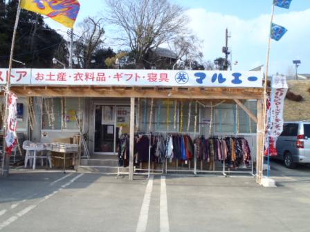 駐車場に面した小さな店舗の前に衣料品が並べられていて、建物の上部には「お土産・衣料品・ギフト・寝具 マルエー」と書かれた看板が掲げられている店の外観写真