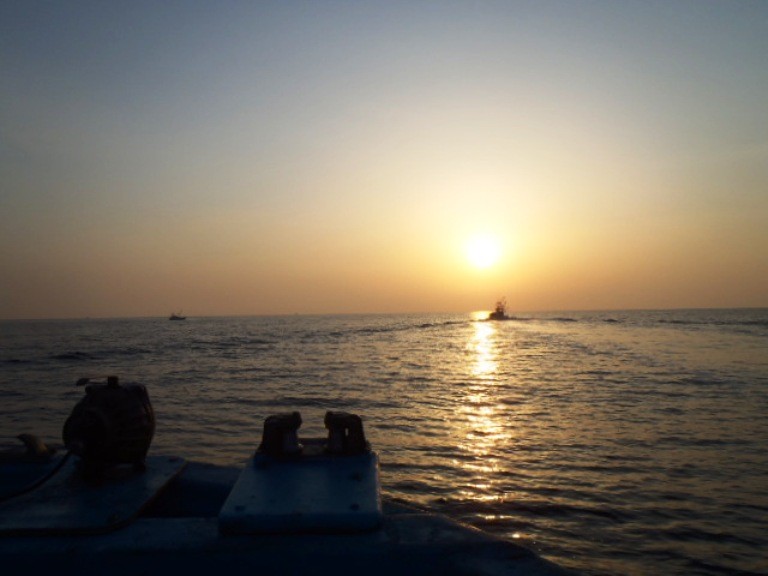 海の上に太陽の光がまっすぐ金色の帯のように伸び、水平線の近くに浮かんでいる一隻の漁船のシルエットが黒く浮かび上がっている写真