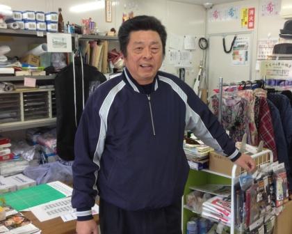 紺色のジャージを着た店主が、商品が並んでいる店舗内で笑顔で写っている写真