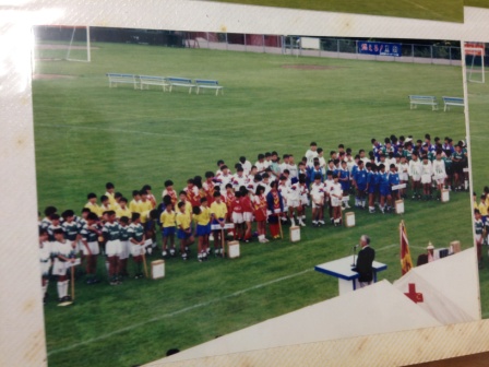 緑の芝生のグラウンドにユニフォームを着た少年サッカーの各チームが整列し、表彰台の前で大会関係者の話を聞いている式の様子を写した、少年サッカー大会の写真