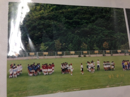 ニフォームを着た少年サッカーの選手たちがいくつかのグループに分かれて芝生のグラウンド上に整列し、フェンスには二つ折りにされた茶色の看板が取り付けられている様子の写真