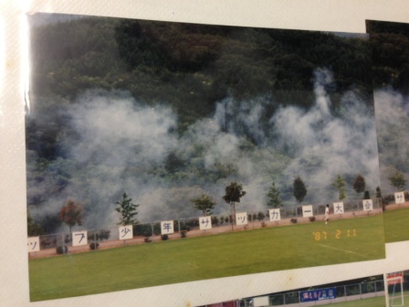 フェンスに取り付けられた看板に仕込まれた導火線に火がつけられ、煙が立ち上る中で自動的に看板が開いて「少年サッカー大会」などの文字が現れる演出の瞬間を捉えた写真。