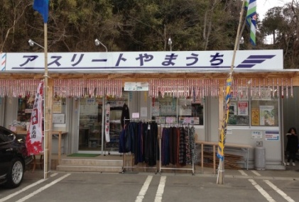 白地に青い文字で「アスリートやまうち」と書かれた看板が掲げられ、のぼりや商品が店頭に並べられている簡易店舗風の建物の正面を写した、スポーツ用品店「アスリートやまうち」の外観写真
