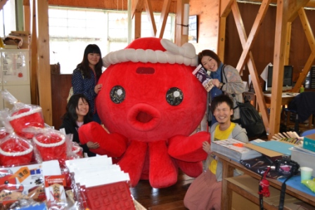 赤いタコの着ぐるみ(オクトパス君)のキャラクターが中央に立ち、その周囲で数人の人々が笑顔で並んで記念撮影をしている様子が写っている写真