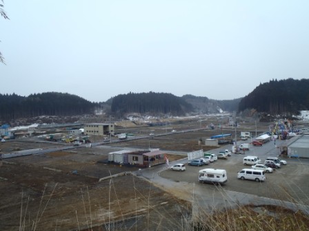 遠くには木々に覆われた山々が見え、広い空き地にいくつかの建物と車が駐車されている様子が見える風景写真