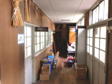 木の板張りの壁に囲まれた廊下の両側にガラス戸の部屋が並び、突き当たりには棚や荷物が置かれている写真